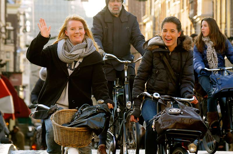 Two young ladies riding bikes in a city, one of them waving at the camera.